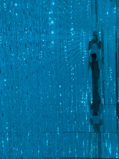 A person is lying down in a room filled with blue lights and mirrors. The person is wearing a white shirt and a white cap, and appears to be lying on their back. Their reflection is visible in the mirrors, creating an illusion of depth and repetition. The room is dark except for the numerous small blue lights scattered throughout, which are also reflected in the mirrors, giving the impression of a starry sky or a digital landscape. The overall atmosphere is serene and immersive. The image appears to be taken indoors, in a controlled environment. There is no indication of the time of day, weather, or specific location beyond the general context provided. No text is visible in the image.
