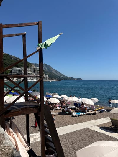 This image depicts a sunny beach scene in Rafailovići, Montenegro. In the foreground, a dark brown wooden multi-level structure, resembling an observation or lifeguard tower, occupies the left side. A light green piece of fabric, possibly a towel or flag, hangs draped over an outward-extending wooden beam on the upper right side of this structure, positioned against the clear blue sky. Below the structure's main platform, several folded beige beach umbrellas are stored. A partial view of a gray concrete wall is visible in the bottom left.
The beach itself is composed of dark gray pebbles or coarse sand. Numerous white beach umbrellas, along with a few colored ones (blue, green, gray), are set up across the midground, accompanied by lounge chairs. Many individuals are present, engaging in typical beach activities; some are reclining under umbrellas, others are standing near the water's edge. A small child in a striped shirt is visible near the stairs of the wooden structure. White slatted wooden pathways traverse the pebble beach. A red trash can is partially visible near the wooden structure, and a yellow inflatable object lies on the sand near the water.
The calm, deep blue-green sea extends across the middle and right sections of the image. A line of orange buoys marks a designated swimming area, where several people are seen swimming or wading. In the background, lush green mountains rise steeply, dotted with numerous white multi-story buildings, likely hotels or apartments, featuring balconies. The coastline recedes to the right, showing a distant headland. The sky is uniformly clear and bright blue.