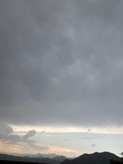 The image depicts a dramatic sky at sunset over a mountainous landscape. The sky is dominated by dark, heavy clouds on the left, suggesting an approaching storm or the remnants of one. As the eye moves to the right, the clouds become lighter and more diffused, transitioning into a bright, hazy band of light at the horizon. This band is illuminated by the setting sun, casting warm, orange and pink hues across the sky and reflecting off some of the scattered clouds.
Silhouetted against the bright horizon are the jagged peaks of mountains, creating a stark contrast between the darkness of the peaks and the vibrant sky. The overall scene conveys a sense of atmospheric drama and the transition from day to night, with a hint of impending weather. The location cues of Dobrsko selo, Montenegro, suggest a rural or mountainous setting. The time of day is clearly sunset, indicated by the colors and lighting. The weather appears changeable, with dark clouds suggesting rain or a storm, contrasted with the clear, illuminated horizon. There are no people or visible objects in the foreground, focusing the attention entirely on the natural elements of the sky and mountains. No text is visible in the image.