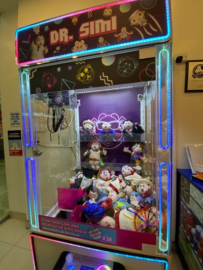 The image shows a Dr. Simi claw machine, a type of arcade game, likely located in San Bernardino Tlaxcalancingo, Mexico. The machine is filled with plush Dr. Simi dolls, the mascot of a Mexican pharmaceutical chain. The machine's signage indicates that it costs 30 pesos to play and that it accepts "Similandia" coins, which can be purchased at the counter. The claw mechanism is visible inside the machine. The exterior of the machine features "DR. SIMI" branding and colorful light strips. Additional shelves stocked with products are visible above the machine. The area appears to be a brightly lit retail space. Signage indicating pharmacy hours and personnel requests are visible on the machine.