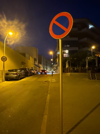 The image shows a street scene at night in Blanes, Spain. A street light illuminates a row of parked cars on the left side of the road. A "No Parking" sign is visible on a pole on the right side of the road. The street is paved with asphalt and there are buildings on either side of the street. The sky is dark, indicating it is nighttime.