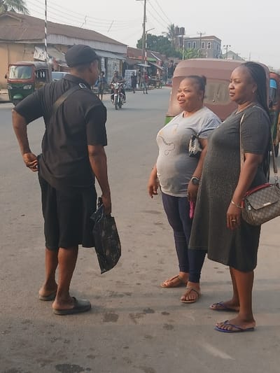 The image displays three individuals on a paved road in Aba, Nigeria, engaged in a conversation under daylight conditions. On the left, a male is seen from behind, wearing a black cap, black short-sleeved shirt, black shorts, and dark slip-on shoes, with a dark strap across his chest and holding a dark, patterned bag in his right hand. He faces two females standing to his right. The first female, positioned centrally, wears a light grey t-shirt with a dark graphic, dark blue leggings, and light brown flat sandals; she exhibits a slight smile. The second female, on the far right, wears a dark grey knee-length dress and blue flip-flops, carrying a brown and beige patterned crossbody bag on her right shoulder; her expression is neutral.
The background reveals a street lined with various commercial and residential buildings, including single-story structures with corrugated metal roofs and taller buildings in the distance. Utility poles with overhead wires are present. Vehicles include a green and red motorized rickshaw (keke Napep) on the far left, another brown motorized rickshaw partially visible behind the women, and a person riding a motorcycle further down the street. The road surface is asphalt with visible stains or patches.
