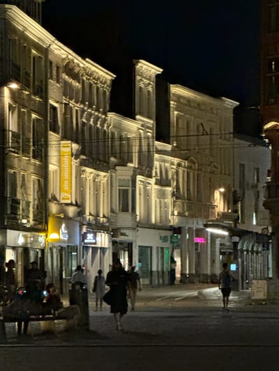 The image depicts a street scene at night in Ghent, Belgium, possibly near the Veldstraat shopping area, known for its historical architecture and shops. The focus is on the illuminated facades of buildings lining the street. A variety of shops are visible, with some displaying bright signage, including a yellow banner with text. Pedestrians are present on the sidewalk, some walking and others seated on a bench. The lighting suggests a commercial area, with streetlights and shop lights casting a warm glow on the scene. The architecture appears to be a mix of older and more modern styles. The overall atmosphere is calm and urban.