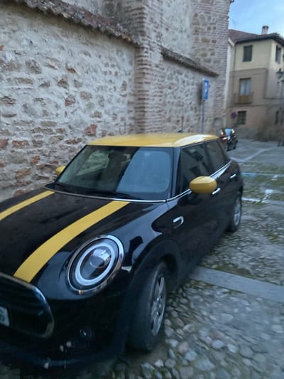 The image depicts a black Mini Cooper with a yellow racing stripe parked against a stone wall in the city of Segovia, Spain, at coordinates 40.952, -4.125. The car is facing right and is parked next to an older building with a stone facade. The car's headlights are on and the windshield is tinted. The car is in focus while the background is blurred. The cobblestone street in front of the car is visible in the foreground. The car is parked on the side of a narrow street that appears to be cobblestone, with some faded paint markings visible on the street. There is an older building visible on the right side of the image.
