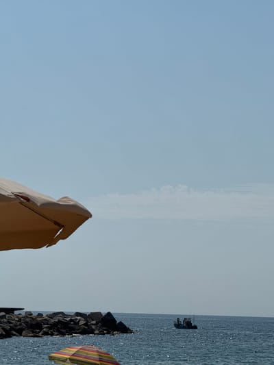 A beach scene in Barcelona, Spain, with a clear, pale blue sky dominating the upper portion of the frame. In the foreground, a portion of a large, light-colored beach umbrella is visible on the left, casting a shadow. Below this, a brightly colored, striped umbrella is partially submerged in the shimmering blue water. A breakwater made of large, dark rocks extends into the sea from the left, forming a barrier. In the middle distance, a small fishing boat with at least two people on board is navigating the water. The sea is calm, with sunlight reflecting off its surface. The overall impression is a peaceful, sunny day at the coast.