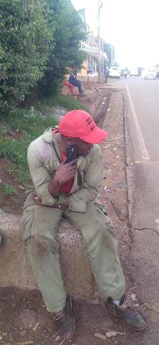 A man in green clothing and a red cap is sitting on the edge of a curb talking on a black smartphone. He is wearing dark brown shoes and a black bag strapped across his chest. He is angled toward the camera with a green bush to his left. In the background are other people walking along the side of the street with vehicles parked.
