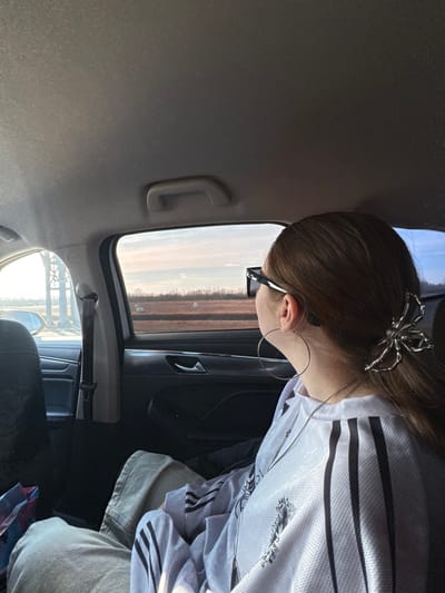 An image captured from within the rear passenger compartment of a vehicle in Verevo, Russia, depicts an individual with brown hair, secured by a silver spider-shaped hair claw, seated and looking out the right-side window. The person wears dark sunglasses, large silver hoop earrings, and a light-colored long-sleeved top featuring three dark stripes on the sleeves. The exterior view through the window reveals a vast, flat, reddish-brown field stretching under a bright, light blue sky with soft pink and orange hues. Several white, possibly spherical objects, are scattered across the field, which is bordered by a distant line of dark vegetation. Through the front driver's side window on the left, a large, light-colored truss structure or tower is partially visible. The vehicle's interior includes a light grey headliner with a grab handle, dark door panels, and a multi-colored plastic bag (blue, pink, purple) positioned on the floor or seat to the left of the individual.