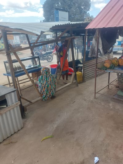 This image captures a street-side market scene in Dar es Salaam, Tanzania. A wooden stall with a corrugated metal roof is visible, displaying pineapples and papayas on one table. Inside the stall, a person wearing an orange shirt and another in a colorful patterned garment attend to business. A glass display case is set up within the stall, possibly showcasing goods for sale. A sign for "Seventh Day Adventist Church" in Kiswahili is seen in the background. Motorbikes and cars pass by on the road outside the stall, indicating a busy street. A few items are scattered on the ground near the stall. The ground is dusty with a single leaf nearby.