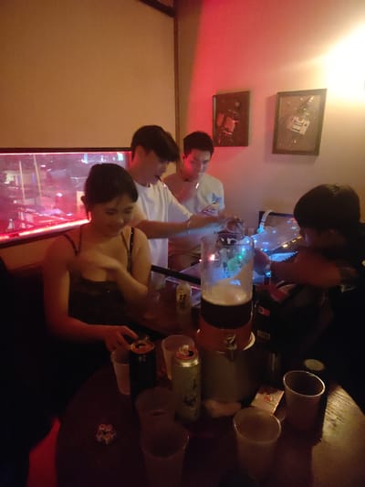 In Nanhai District, China, at what appears to be a bar or restaurant, four young adults are gathered around a table. A woman in a dark, strappy top is seated on the left, holding a cup and looking down. To her right, a man in a white t-shirt is standing, talking to another man who is also in a white t-shirt and is partially obscured. The third man is on the far right, facing away from the camera and appearing to interact with a device. In the center of the table is a large beer dispenser with illuminated lights, surrounded by several cups and two beer cans. The environment is dimly lit with red and yellow lighting, and two framed pictures hang on the wall in the background.