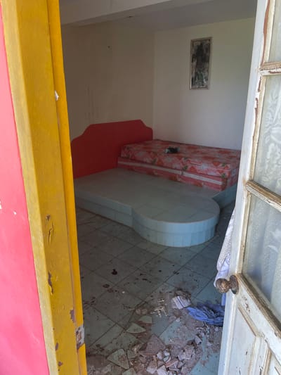 The image depicts a disheveled room, possibly in Phu Thuy Ward, Vietnam, with a focus on a raised sleeping platform covered by a floral-patterned mattress. The room appears to be in a state of disrepair. The floor is tiled and littered with debris, including broken pieces of what looks like concrete or plaster, and some crumpled fabric. A red headboard is attached to the wall behind the mattress. To the right, a partially open wooden door with peeling paint and a tarnished doorknob is visible, revealing a glimpse of an exterior or another room. A framed picture hangs on the wall above the bed. The framing of the doorway to the left is brightly painted in pink and yellow. The overall impression is one of neglect and disarray.