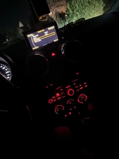 The image shows the interior of a car at night. The dashboard is illuminated by red lights from the radio and climate control system. A small screen displays "Radio FM" and "Waveband" at the top, with radio station information below, including "105.8 FM FOLK" and "87.8 MHz 107.7 MHz". A speedometer is visible on the left, with a needle pointing between 140 and 160. Outside the car, trees and buildings are faintly visible in the darkness. There is no indication of the specific location of Karnobat, Bulgaria within the image. No people are visible in the image.