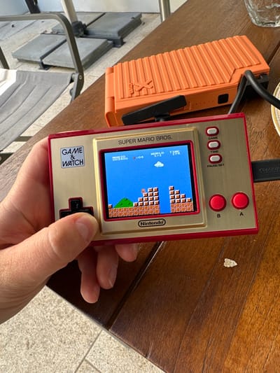 An outdoor scene in Vandœuvres, Switzerland, depicts a person's left hand holding a powered-on Nintendo Game & Watch: Super Mario Bros. handheld console. The device, gold with red accents, displays "WORLD 1-1" of the game with the pixelated Mario character positioned on brick platforms against a blue sky. The individual's thumb rests on the console's black directional pad. A black cable is plugged into the console's right side, extending towards an orange, ridged portable electronic device, likely a power bank, which rests on the dark wooden table. In the blurred background, grey outdoor furniture components and light-colored patio pavers are visible under natural light. A small, light-colored crumb is present on the table surface to the right of the gaming device.