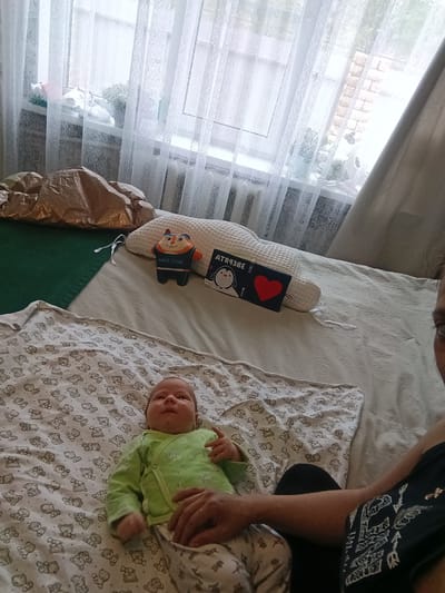 A baby wearing a light green long-sleeved shirt and white patterned pants lies on a patterned blanket. The blanket is spread on a light grey bed, with a woman's hand touching the baby's stomach. To the baby's right are two toys and a small book with a penguin illustration and the Russian word "Зверюшки" printed on it, next to a heart. Behind the baby, more objects are on the bed: a brown pillow-like object on a green surface and a white pillow. A window covered with sheer white curtains is in the background. The scene is likely indoors in Andreyevo, Russia, though this cannot be determined definitively from the image.