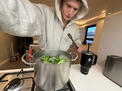 This image captures a rotated, medium close-up view of a person cooking broccoli in a kitchen. The individual, appearing male with fair skin and light hair, is wearing a light grey zippered hooded sweatshirt and a silver chain necklace, and looks directly at the camera with a neutral expression. They hold a pair of metal tongs in their left hand.
The main activity shows a large, silver metal pot on a gas stovetop, where green broccoli florets are actively boiling in water, with visible steam and bubbles. To the left of the pot, on a white countertop, stands a black Hydro Flask coffee mug with a handle. The mug prominently displays the white Hydro Flask logo (a stylized 'X' within a circle), and the text "Hydro Flask" is visible on its lid.
The kitchen setting includes white countertops and what appears to be a gas stovetop with black grates. A silver-toned appliance is partially visible on the far left. In the background, through a window, a bright blue sky and some distant structures suggest it is daytime. Recessed lighting is visible on what would be the ceiling or under cabinets. The entire image is rotated approximately 90 degrees counter-clockwise, giving an inverted perspective of the scene. The location is specified as London, United Kingdom.