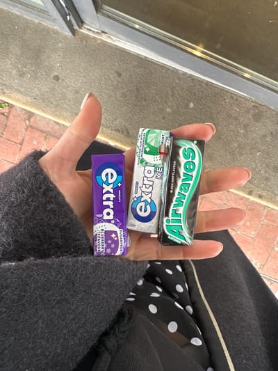 A human left hand is centrally positioned, displaying three packets of chewing gum. The hand exhibits short fingernails with a light, possibly nude-colored polish, showing minor wear at the tips. The gum packets, from left to right, are: a purple "Extra" brand packet, identified as "BLUEBERRY FLAVOUR" and "SUGARFREE"; a white and green "Extra ICE" packet, indicated as "MINT" and "SUGARFREE GUM WITH MICROGRANULES"; and a black and green "Airwaves" packet, labeled "BLACK MINT FLAVOUR" and "WRIGLEY'S".
The individual's dark grey, textured sleeve is visible on the left side of the frame, covering part of the forearm. Below the hand, a black garment with white polka dots is discernible, adjacent to a plain dark fabric segment featuring a vertical, thin, light-colored stripe.
The background consists of an urban outdoor setting. In the foreground, a rough grey concrete surface transitions to a section of red brick paving, laid in a stretcher bond pattern. Above this, a dark-framed window or door with reflective glass is visible, suggesting a building facade. The precise location within Tunbridge Wells, United Kingdom, cannot be determined from the available visual information, but the environment is consistent with a public street or pedestrian area.