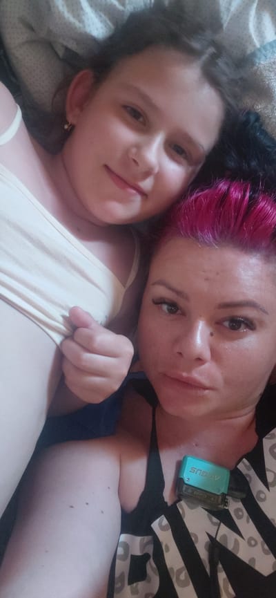 Two people, likely an adult and a child, are lying down close together, looking up at the camera. They are in a horizontal orientation. The person in the foreground has bright pink hair, dark eyes, and is wearing a black top with a white patterned design. A turquoise object, possibly a vape or electronic device, is visible on their chest, with the word "ARGUS" printed on it. The person in the background has lighter skin and dark hair. They are wearing a light-colored top. The setting appears to be indoors, possibly a bed, given the soft bedding visible. The lighting suggests it might be daytime, but there are no strong indicators of the time of day or weather conditions. The overall mood seems relaxed and intimate.