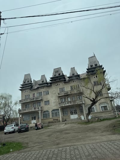 The scene depicts a street view in Jilava, Romania, under an overcast, light grey sky. A multi-story building with a weathered, light grey facade dominates the midground. This structure features ornate architecture with at least five prominent, steep, grey-tiled Mansard-style roofs, each capped with decorative finials. Multiple windows with dark frames and balconies with concrete railings are visible on the upper floors. Decorative elements, including patterned frames, adorn the windows and sections of the facade. The ground floor includes large windows and a central entrance with columns supporting an upper balcony.
In the immediate foreground, a dark grey patterned paved surface is visible, transitioning to a dirt and gravel area. Parked on this unpaved surface are three vehicles: a white Dacia Logan sedan (license plate IF 97 VLF), a black Opel Astra hatchback (license plate IF 18 PKO), and a three-wheeled vehicle covered with a tan tarp. Two individuals are partially visible near the white sedan; one in dark attire is near the front driver's side, and another in lighter clothing is positioned near the rear.
A large, deciduous tree with a thick, gnarled trunk and visible green foliage from new growth stands in the mid-right, partially obscuring the building. Another bare-branched tree is visible on the far left. Overhead, multiple dark electrical power lines stretch across the upper portion of the frame, originating from a utility pole partially visible on the far left.
