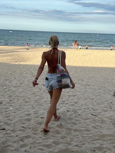 A woman with blonde hair styled in a braid walks away from the camera on a sandy beach. She is wearing a brown bikini top, light blue denim shorts, and flip-flops. She carries a patterned tote bag over her left shoulder. A cell phone is visible in the back pocket of her shorts. The beach is sparsely populated with a few other people in the distance, some in the water, some on the shore. The ocean is visible behind the beach, with a clear sky above. The image appears to have been taken at a beach in Da Nang, Vietnam.