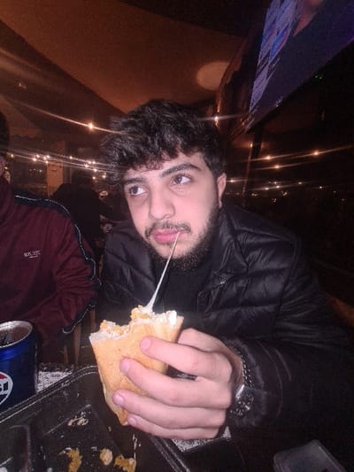 A man in a black puffer jacket eats a sandwich with stringy cheese, tongue out. He wears a silver watch on his left wrist. A dark metal tray beside him holds the sandwich. A partially visible beer can sits near the tray's edge. Soft lights are visible behind the man. He has dark hair and a beard. A television screen is visible behind him. Part of another person is visible wearing a red shirt.