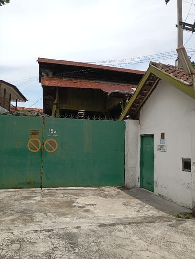 The image shows a green metal gate with a sign that reads "15 A" and "L.21/B11," two yellow "No Parking" signs, and a white building to the right of the gate. The building has a green door and a small window. The gate has barbed wire and a wooden frame. There is a rusted, corrugated metal roof in the background, which is partially obscured by the white building. The scene is in Katapang, Indonesia, located at the coordinates latitude -6.985, longitude 107.56. The ground is concrete. The image is taken from the street in front of the gate, looking at the gate and the building to the right. The sky is cloudy. There are no people visible in the image.