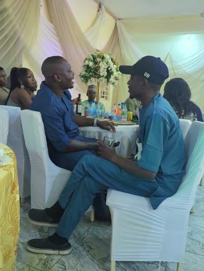 Two men in matching blue traditional attire are seated at a table during a gathering in Barden Yana, Nigeria. The man on the left is facing the man on the right, with his mouth open as if speaking. He is holding a clear glass in his right hand. The man on the right is wearing a navy blue baseball cap with "GB" on it and is looking towards the man on the left. The table is covered with a white tablecloth and has several bottles of beverages and a floral centerpiece. In the background, other guests are visible, and the area is decorated with draped white fabric, suggesting an indoor event.