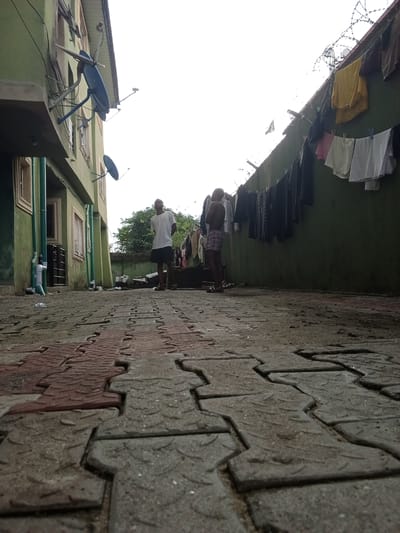 In Ado, Nigeria, an outdoor scene unfolds on a paved courtyard between two buildings, with one to the left and another to the right, both painted in a muted green hue. Laundry hangs on a clothesline stretched along the side of the building on the right, with barbed wire visible above it. Two young males are present in the courtyard. One stands near the left building, wearing a white t-shirt and dark shorts. The second young male is positioned further back, closer to the right building, and is shirtless, wearing checkered shorts. The foreground is dominated by interlocking paving stones, some red and some gray, with a textured surface. The lighting suggests an overcast sky, casting a diffused light on the scene.
