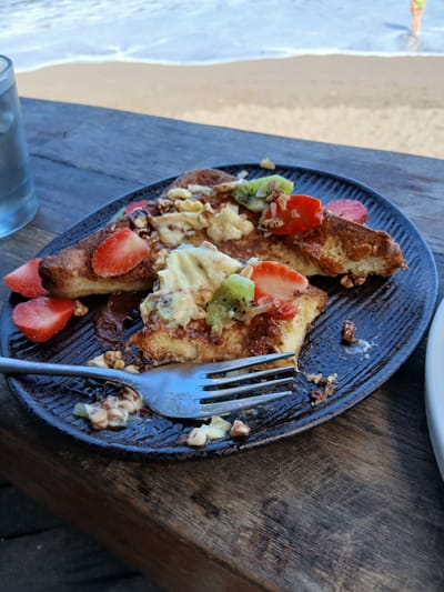 A plate of French toast with strawberries and kiwi fruit sits on a wooden table by the ocean in an unknown location in Indonesia. The plate is dark and textured with a wavy pattern. The French toast appears to be topped with a creamy sauce, possibly whipped cream or custard, and chopped nuts. Slices of strawberry and kiwi are scattered around the toast. A fork rests on the plate, ready to take another bite. The ocean waves can be seen in the background, with a sandy beach stretching out before them. The overall impression is one of a relaxing and delicious meal enjoyed in a beautiful setting.