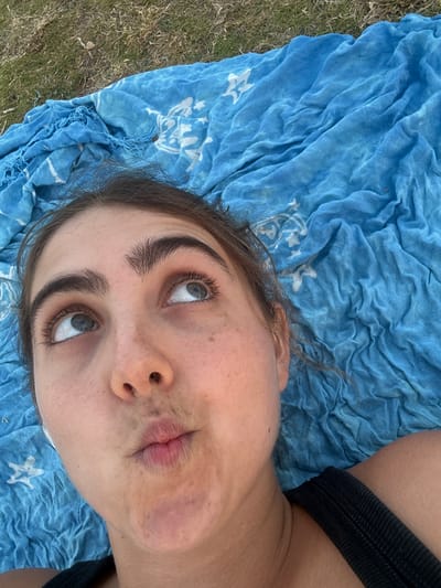 A young woman is lying on a blue towel on grass, looking up and to the right with a pouty expression. She has dark eyebrows and eyelashes, and her eyes are visible. The towel is draped behind her head and shoulders, and appears to be made of a lightweight fabric with a pattern of white stars. The background is out of focus, showing grass and possibly other foliage. The woman is wearing a black strap, possibly from a tank top. The lighting suggests an outdoor setting during the day.