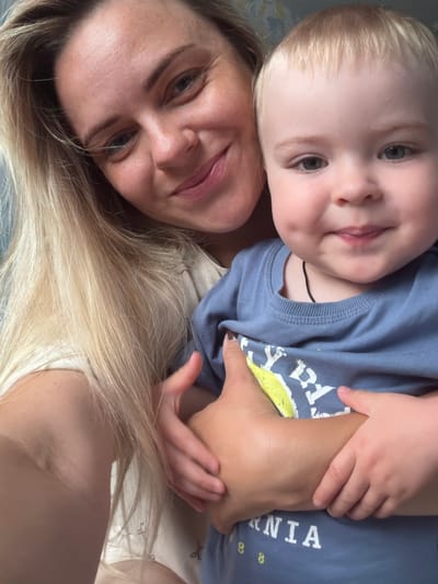 This is a close-up, selfie-style image of a woman and a young child. The woman, with blonde hair and fair skin, is looking towards the camera with a slight smile. Her face is in focus in the foreground. The child, with blonde hair and blue eyes, is positioned behind and slightly above the woman, with their head resting on her shoulder. The child is looking slightly away from the camera.

Both subjects are wearing casual clothing. The child is wearing a blue t-shirt with yellow lettering on the front and a thin black cord around their neck. The woman is wearing a light-colored top, possibly a t-shirt or tank top. Their hands are intertwined, with the child's hand resting on the woman's arm.

The overall mood of the image appears warm and affectionate, suggesting a close bond between the two. The lighting is soft, indicating it could be daytime, though no specific time is discernible. There are no clear environmental cues to suggest the specific location within Votkinsk, Russia, or the weather conditions.

The visible text on the child's shirt appears to be part of a word, possibly "NIA" is visible, along with some smaller yellow markings that resemble numbers or symbols. The image is oriented vertically.