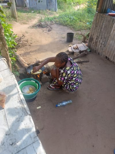 A woman with braided hair, wearing a colorful patterned dress, is crouched on the ground outdoors, cooking. She is tending to food in a black pan over an open fire, which is made from wood and stones. Next to her is a stack of green and white buckets containing some food items, and a metal colander. The ground is sandy and uneven, with some sparse vegetation and debris scattered around. In the background, there are corrugated metal structures and wooden posts. A plastic water bottle lies on the ground in front of her, and a white table with some plastic bags is visible on the left side of the image. The scene appears to be in a rural or informal settlement area, likely in Dar es Salaam, Tanzania, given the context.