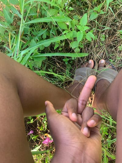 A low-angle shot focuses on a person's feet in sandals, standing on grass and dirt. One hand gestures with an extended middle finger. The background is lush green foliage and some small purple flowers. The person's brown skin tone contrasts with the natural environment.