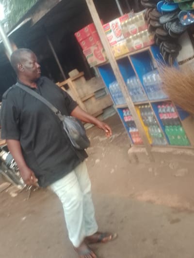 An outdoor scene in Alor, Nigeria, features an adult male positioned slightly left of center, facing towards the right. He has dark skin and short dark hair, wearing a black short-sleeved shirt, light-colored trousers, and dark sandals. A dark shoulder bag is worn across his chest. His right arm is slightly extended forward and down. Behind him, a retail stall is visible, constructed with wooden elements and blue shelving. The shelves display various goods: the top sections hold stacked cardboard boxes, some labeled "Indomie," alongside bottles of beverages, including some with "Amstel" branding. The middle shelves contain rows of clear plastic water bottles. The bottom shelves hold an assortment of plastic bottles containing different colored soft drinks. To the immediate right of the shelving unit, a vertical stack of numerous flip-flops or sandals, including blue and black pairs, is present. The ground surface is unpaved earth. A segment of a straw broom extends into the frame from the extreme right foreground. A metallic object, possibly a part of a bicycle or motorcycle, is partially visible behind the man on the left.