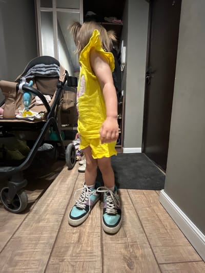 The image shows a young child standing in what appears to be a hallway or entryway of a home. The child is wearing a bright yellow dress and colorful sneakers. They are positioned in the center of the frame, with their back mostly to the camera. To the right of the child, a portion of a stroller is visible, suggesting the child may have been using it or is about to. The floor is made of light-colored wood planks. In the background, a dark doorway or closet is visible, along with shelves containing various items. The lighting suggests it might be daytime, but the interior setting makes it difficult to determine the exact time of day or weather conditions. There is no visible text in the image. The overall scene conveys a sense of domesticity and everyday life.