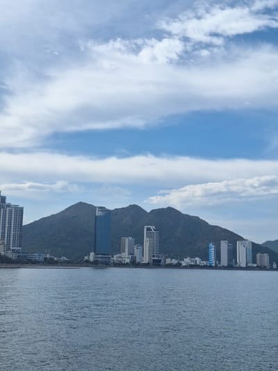 A panoramic view shows the coastline of Nha Trang, Vietnam, featuring a body of water in the foreground and a city skyline backed by lush green mountains under a cloudy sky. The water is a deep blue with subtle ripples. The city skyline is composed of numerous modern high-rise buildings of varying heights and designs, predominantly white and grey, with a few featuring blue facades. A distinct tall, slender building with a blue exterior stands out amongst the others. Behind the city, a large, mountainous landmass covered in dense green vegetation dominates the background, its peaks softened by distance and atmospheric haze. The sky is a mix of blue and white, with streaky clouds creating a dynamic pattern. There are no discernible people or actions depicted in the image.