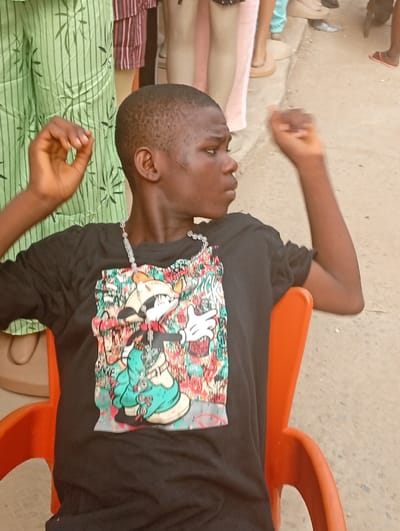 An adolescent male is seated in an orange plastic chair within an outdoor market setting in Nkwelle, Nigeria. He has a dark complexion and short, dark hair. He is wearing a black t-shirt adorned with a large, colorful graffiti-style graphic featuring a cartoon character and a translucent beaded necklace. His arms are slightly raised, bent at the elbows, with both hands open. He is looking towards the right side of the frame with a neutral expression. The background reveals several headless mannequins displaying various garments, including green patterned trousers, striped clothing, and light-colored pants, indicative of a clothing retail stall or shop. The ground surface is light-colored and appears unpaved.