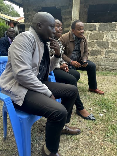 Four individuals are captured in an outdoor setting, seated on blue plastic chairs against a backdrop of a rough-hewn stone wall and grassy ground. The location is Kiganjo, Kenya.

In the foreground, from left to right, are three individuals. The leftmost person is a bald man wearing a light grey jacket over a dark, patterned shirt and black trousers. He sits with his left hand near his chin, seemingly deep in thought or engaged in conversation. Next to him is a woman in a black and white animal-print top, black trousers, and black flat shoes with a bow detail. Her hands are clasped in her lap, and a colorful beaded bracelet adorns her wrist. She appears attentive. To her right is a man wearing a brown jacket over a dark patterned shirt and black trousers, sporting brown dress shoes. He is holding a mobile phone in his left hand and wears rings on both hands. All three are looking towards the right side of the frame, suggesting they are observing an event or person outside the camera's view.

Further in the background, partially obscured behind the man in the grey jacket, another man in a dark jacket is visible, also seated on a blue plastic chair. The ground is dry and grassy, with some scattered debris. The natural lighting indicates it is daytime, likely a bright or sunny day. No text is visible in the image. The scene suggests a casual outdoor gathering or meeting.
