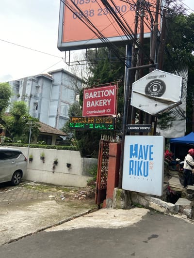 The scene is located in Bandung, Indonesia, at coordinates -6.88 latitude and 107.603 longitude. It shows a street with a gray car parked on the left, facing a white wall with a gate. On the right side of the image, a black pole is attached to several signs. The top sign is white with a circular red logo for "Laundry Shoes" and the text "The Lazy." Below it is a red sign that reads "Bariton Bakery Clumbuleuit 101." Below this is a green digital sign displaying a price list. The bottom sign is blue and white and reads "Mave Riku Coffee." There is a blue canopy in the background and a person in a red jacket standing near a motorcycle. The environment includes a concrete parking lot, a wall with a metal gate, and a sidewalk in front of a coffee shop. There is debris scattered on the ground.