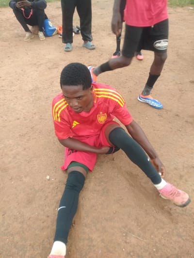 The image shows a group of people, likely a soccer team, on a dirt field in Durumi, Nigeria. The main subject is a young man in the foreground, wearing a red and yellow soccer jersey, black shorts with green accents, and black shin guards. He is sitting on the ground with his legs extended, appearing to be resting or tending to his equipment. His expression is focused and somewhat tired.
In the background, other individuals are visible, also dressed in athletic attire. One person is crouching and holding a phone. Another person's legs are visible, wearing black shorts and blue and orange soccer cleats. A blue bag with "Realme" written on it is on the ground near the crouching person. The ground is a reddish-brown dirt, characteristic of many outdoor fields in Nigeria. The lighting suggests it might be daytime, possibly late afternoon, with soft shadows. The overall scene conveys a sense of casual athletic activity, possibly a practice or a break during a game. There is no visible text other than on the bag.