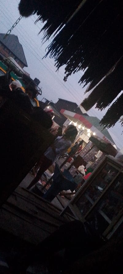 The image presents a tilted, low-light street or market scene in Aba, Nigeria, observed at dusk or night. The upper portion of the frame is dominated by a dark, textured, possibly thatched or frayed awning in the upper right, and a metal chain hanging in the upper left. The sky, visible above the structures, is a dim grey, traversed by power or communication lines.
In the mid-ground, a brightly illuminated market stall or shop is central on the right side, featuring a red and white striped canopy and multiple interior lights. A person, seen from behind, is prominent near the center-right, wearing a light-colored long-sleeved top and dark lower garments, facing towards the lit stall. A blue, possibly plastic, object resembling a container or stool rests on the ground in front of this individual. To the left, several other indistinct figures are visible in darker clothing, some with light-colored head coverings, suggesting a crowded environment. Buildings are discernible in the background against the dark sky. The ground surface is indistinct and dark.
