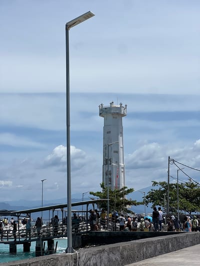 The image depicts a bustling coastal scene in Toyapakeh, Indonesia, under a partly cloudy sky with a distinct horizontal band of haze. In the foreground, a gray concrete pier or seawall extends horizontally, with turquoise water and small waves to its left. A tall, gray metal pole with a solar-powered street light fixture is centrally positioned. To the left, a covered gangway with a dark metal frame and roof extends over the water, supported by multiple pylons. Numerous individuals, appearing to be a crowd of people in various casual attire, are gathered on this gangway and on the adjacent land area, standing and appearing to wait or move. Rocks line the shore beneath the gangway.
In the mid-ground, a tall, white, multi-story tower with a weathered appearance stands prominently. This tower features vertical recesses along its sides, rectangular openings, and a distinctive ornate, crown-like observation deck at its apex with decorative railings. A thin red banner or flag is partially visible hanging from its lower section. Trees with green foliage are present near the base of the tower and along the shoreline. Further in the background, a series of distant mountains or large hills rise under the cloudy sky. Additional street lights with solar panels and utility poles with overhead wires are scattered throughout the mid-ground. The overall environment suggests a busy port or ferry terminal.