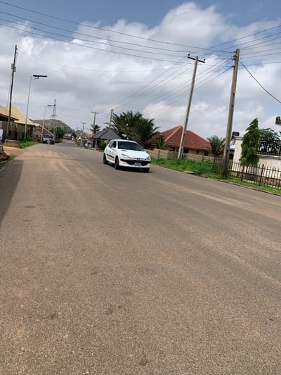 The image captures a slightly overcast day in Barakin Akawo, Nigeria. The scene is a paved road running through a residential area, lined with houses and vegetation. A white Peugeot car is parked on the side of the road, its front partially visible. Telephone poles with numerous wires stretch across the sky, indicating the presence of utilities.

The houses visible have red-tiled roofs and are interspersed with palm trees and other greenery. In the background, a rocky hillside is visible, suggesting the geographical setting. The sky is a mix of blue and grey clouds, indicating potentially changeable weather. No people are clearly visible in the image, though there may be movement within the houses or unseen individuals.

The time of day appears to be daytime, likely mid-morning or afternoon, given the quality of light. The road surface is asphalt, showing some wear and tear. The overall impression is of a typical, somewhat rural or semi-urban, Nigerian street scene. There is some text on the license plate of the car, but it is not legible.