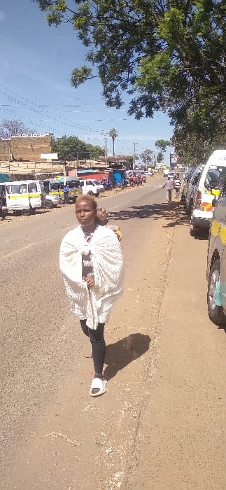 The image depicts a bustling street scene, likely in Nyeri, Kenya, under bright, clear daylight. In the immediate foreground, a person stands on the dusty shoulder of an asphalt road, facing the viewer. They have light-colored hair and are largely enveloped in a distinctive white, textured garment, possibly a shawl or traditional wrap, that extends down to their knees. They are also wearing dark trousers and white sneakers. A prominent, elongated shadow of the person is cast on the ground to their left, suggesting the sun is high in the sky.

Behind the person, the road stretches into the midground, occupied by several vehicles. These include cars and what appear to be Kenyan public transport vans (matatus), some white with yellow and black livery, indicating a busy thoroughfare or parking area. To the left of the road, a row of informal structures, possibly market stalls or small businesses, lines the street, adding to the commercial activity of the area.

The background on the left side of the frame features large, leafy green trees against a clear, light blue sky. Further back, more distant trees and faint structures can be seen. There are also visible power lines or communication cables crisscrossing the sky. The overall scene conveys a lively, active urban or semi-urban environment on a sunny day. No discernible text is clearly visible on any signs or vehicles.