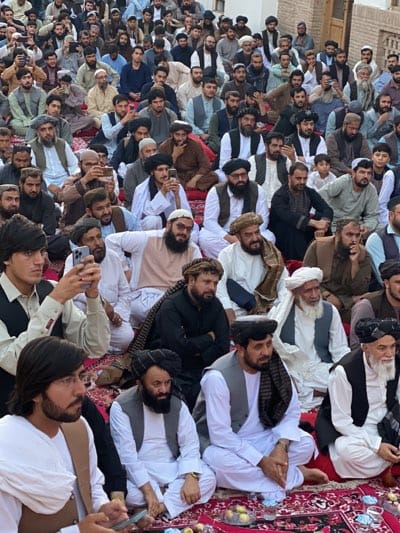 A large group of men are sitting on the ground in an outdoor setting, appearing to be in a public gathering or event. The men are predominantly dressed in traditional Islamic attire, including turbans, shalwar kameez, and vests. Many of them have beards. Some individuals are holding mobile phones, suggesting they are either documenting the event or communicating. The ground is covered with patterned rugs. In the background, there are buildings with architectural features that are consistent with the city of Kandahar, Afghanistan. The overall atmosphere appears to be one of community or religious observance.