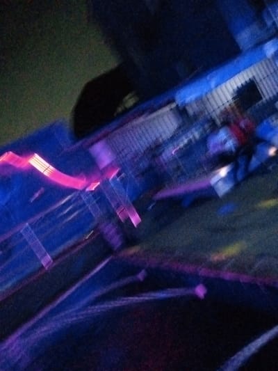 This is a motion-blurred, low-light photograph of an outdoor scene, likely at night. The predominant colors are dark blues and purples, with streaks of bright pink and some hints of yellow and green. The scene appears to be a street or walkway lined with some form of railing or fence, suggested by horizontal and vertical lines that are heavily blurred. There are also diagonal light streaks that indicate movement. In the background, a dark, indistinct shape could be a building or a large object. The overall impression is one of a dynamic, possibly festive or event-like atmosphere due to the bright, streaking lights. The image is too abstract and lacking in clear detail to definitively place it in Ado, Nigeria or any specific location.