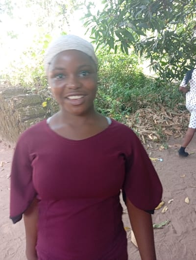 A smiling young woman in a burgundy top and light headscarf stands outdoors. Her complexion is dark. She has dark hair and light eyes. The background features greenery and a stone structure to the left. Another person, partially visible, is wearing a white and black polka dot dress. The ground appears to be covered in dirt or sand. The image is well-lit.