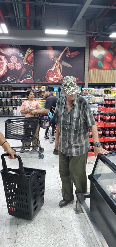 The image captures an indoor scene within a supermarket, likely in Barquisimeto, Venezuela, characterized by bright fluorescent lighting and a terrazzo-tiled floor.

In the foreground, an elderly man wearing a plaid shirt, olive green pants, and a camouflage bucket hat stands intently in front of a glass-fronted refrigerated display case. Glasses are tucked into his shirt, and a wristwatch is visible on his left arm as his hand rests on the display's edge.

Behind the elderly man, a woman with dark hair, dressed in a light pink top, stands beside a black shopping cart with wheels. A man with a beard and a dark cap is partially visible behind her. In the very immediate foreground, a hand holds a black plastic shopping basket, indicating the presence of another shopper (likely the photographer).

The supermarket environment features high ceilings with exposed ducts and bright rectangular lights. On the right, shelves are heavily stocked with numerous red bottles of Coca-Cola, with the brand name clearly visible. The refrigerated display case the man is examining contains various packaged items.

Large, colorful posters adorn the walls, featuring food imagery. One depicts red apples with green leaves and a sliced white apple. Below it, a striking image shows a large bone-in steak, possibly a tomahawk, being cut with a knife. Further down, another poster displays sausages or meat patties alongside another cut of red meat.

The overall scene depicts people engaged in grocery shopping, browsing and selecting items from the store's offerings. The visible text primarily includes "Coca-Cola" on the beverage bottles, along with "Cola," "Coca-C," and "Cocarla" detected from various labels.