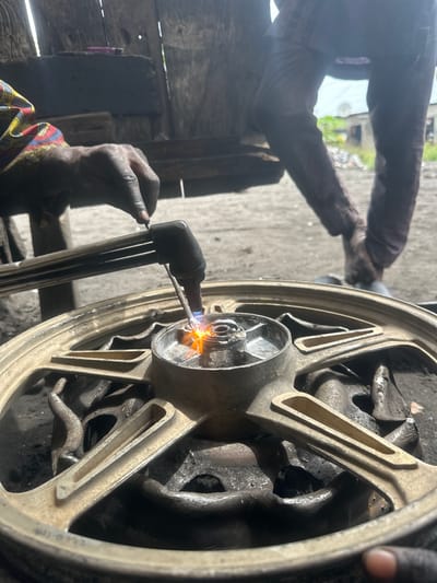 In this medium shot, two individuals are seen in what appears to be a workshop in Badagry, Nigeria. The image is oriented vertically, with the main focus on a gold-colored wheel rim, likely from a motorcycle or a similar vehicle. One person, whose hands are visible, is holding a welding torch, emitting a bright orange flame. The torch is directed at the center hub of the wheel, suggesting a repair or modification is in progress. The individual's hands are dark and appear smudged with grease or dirt, indicative of manual labor. They are wearing a colorful patterned shirt with red, yellow, and blue accents.
In the background, another person's arms and torso are visible. They are wearing a dark long-sleeved shirt and dark pants. Their hands are also dark and appear to be engaged in some work on the wheel, though their exact action is obscured. The setting is dimly lit, with rough wooden structures and concrete flooring, typical of a workshop environment. The overall atmosphere suggests a working scene, with no overt display of emotions. The time of day and weather are not discernible from the image. There is no visible text in the frame.