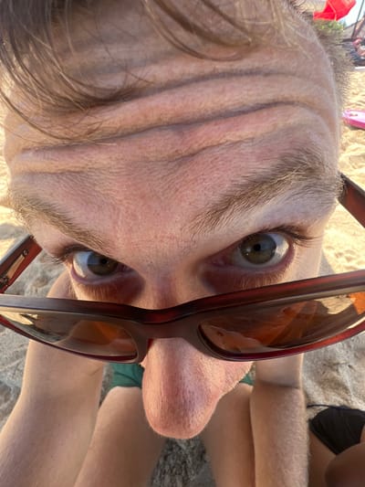 A close-up, low-angle shot shows a person's face from a beach perspective, with a dominant focus on the upper forehead and eyes, framed by sunglasses. The forehead displays prominent horizontal wrinkles. The individual wears sunglasses with dark, reflective lenses that obscure the eyes partially, revealing a portion of the irises. The nose is visible below the sunglasses, and the chin and upper lip area are also in frame. The subject's hair is short and light brown. The background suggests a beach environment, with sand visible in the lower portion of the frame, and elements like a red umbrella and possibly beach chairs or towels in the distance. The scene is lit by natural sunlight, casting a warm glow. The location is Kuta Selatan, Indonesia.