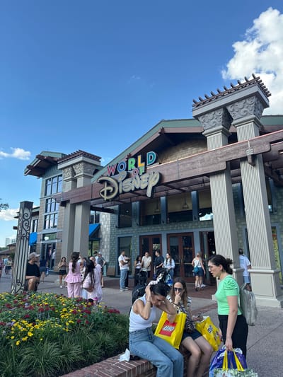 The image shows the World of Disney store in Lake Buena Vista, United States. A large "World of Disney" sign adorns the front of the building. A group of people is sitting on a brick ledge in front of the store. Some of them have shopping bags from Lego and other stores. There are flowerbeds in front of the ledge with a variety of colorful flowers. The sky is blue with some clouds.