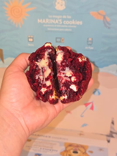 The image displays a person's hand holding a rich, red velvet cookie, split in half to reveal its decadent interior. The cookie is a deep crimson color, generously studded with white chocolate chips, and features a creamy, gooey white filling that oozes from the center, suggesting a soft and moist texture.

The cookie is held against a light blue, illustrated backdrop, which appears to be the packaging for "MARINA'S cookies." The box is adorned with cheerful, summery graphics, including a bright orange sun, a wide-brimmed hat, a small beach umbrella, a beach ball, and a friendly cartoon character resembling a dog or bear. The text "La magia de las MARINA'S cookies" is clearly visible on the box, identifying the brand.

The scene is captured under warm, artificial lighting, which highlights the cookie's vibrant color and the inviting, soft consistency of its filling. The overall impression is one of indulgence and deliciousness. This moment of presenting the appetizing cookie takes place in Santiago, Chile.